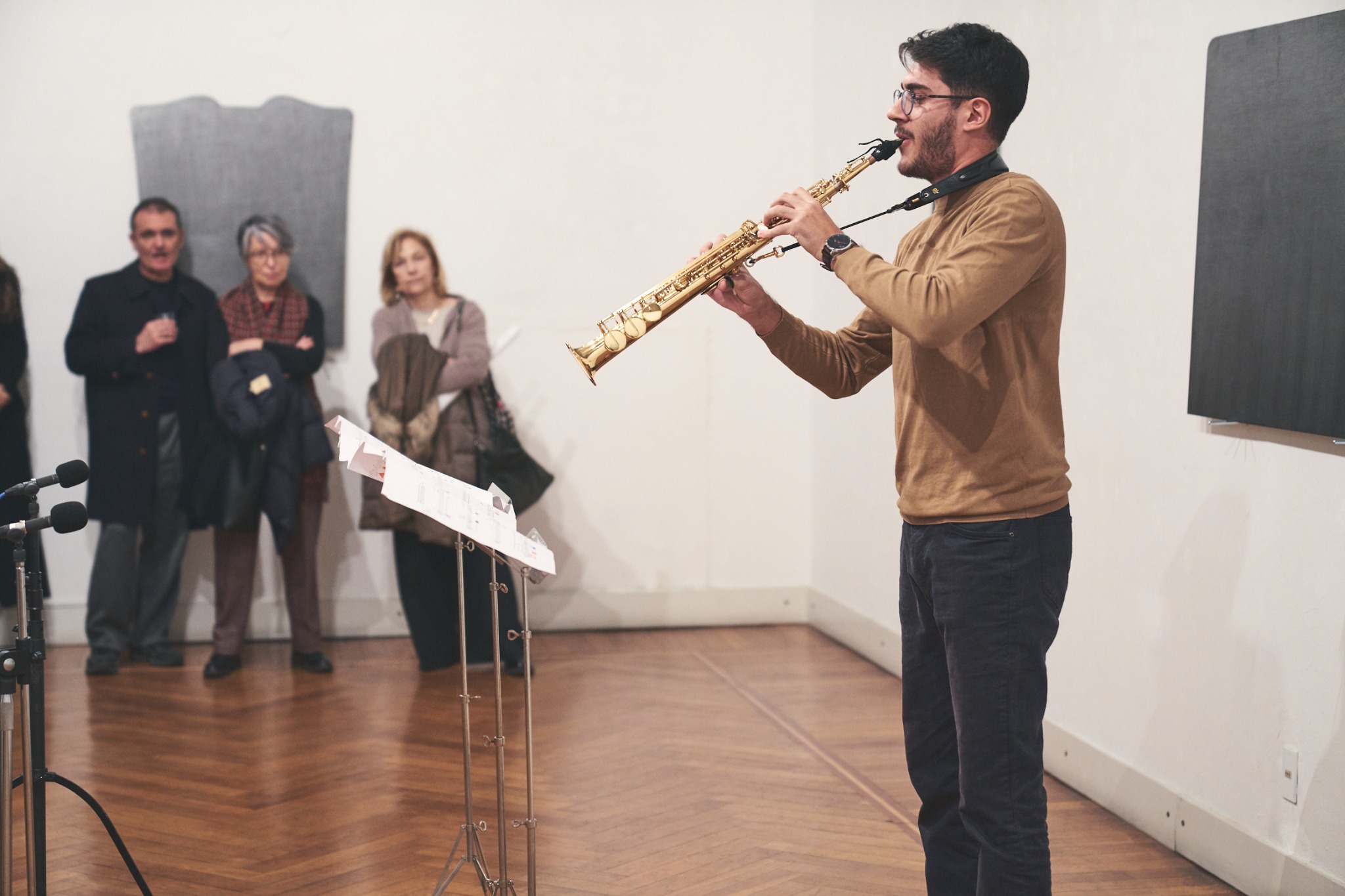 Alberto Cavallaro playing soprano saxophone in a gallery, with listeners nearby.