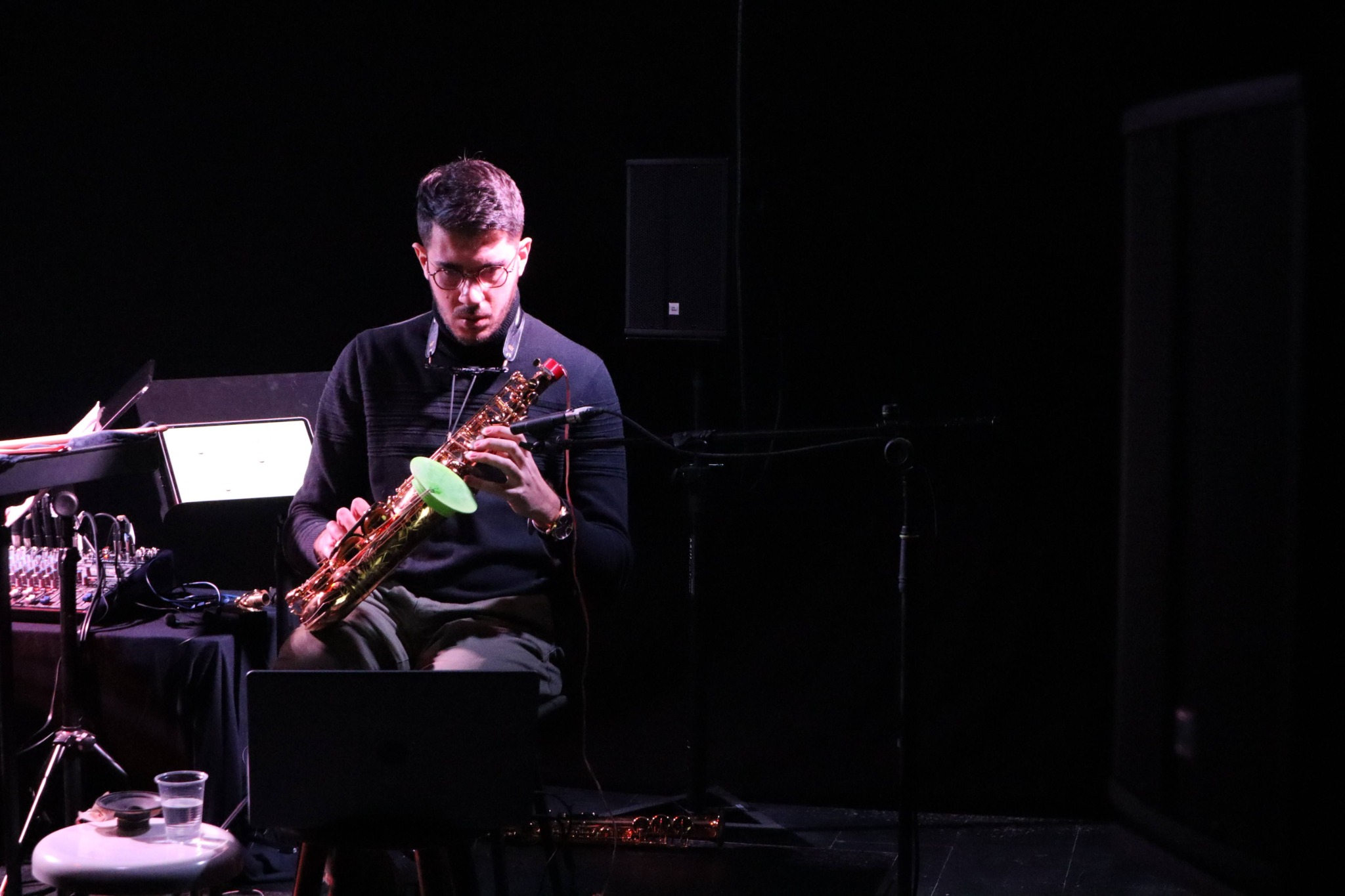 Alberto Cavallaro playing saxophone on a dark stage beside electronic equipment.